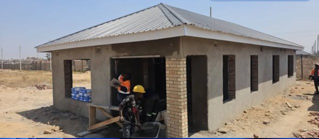 A single-storey house under construction with the roof on and builders at work